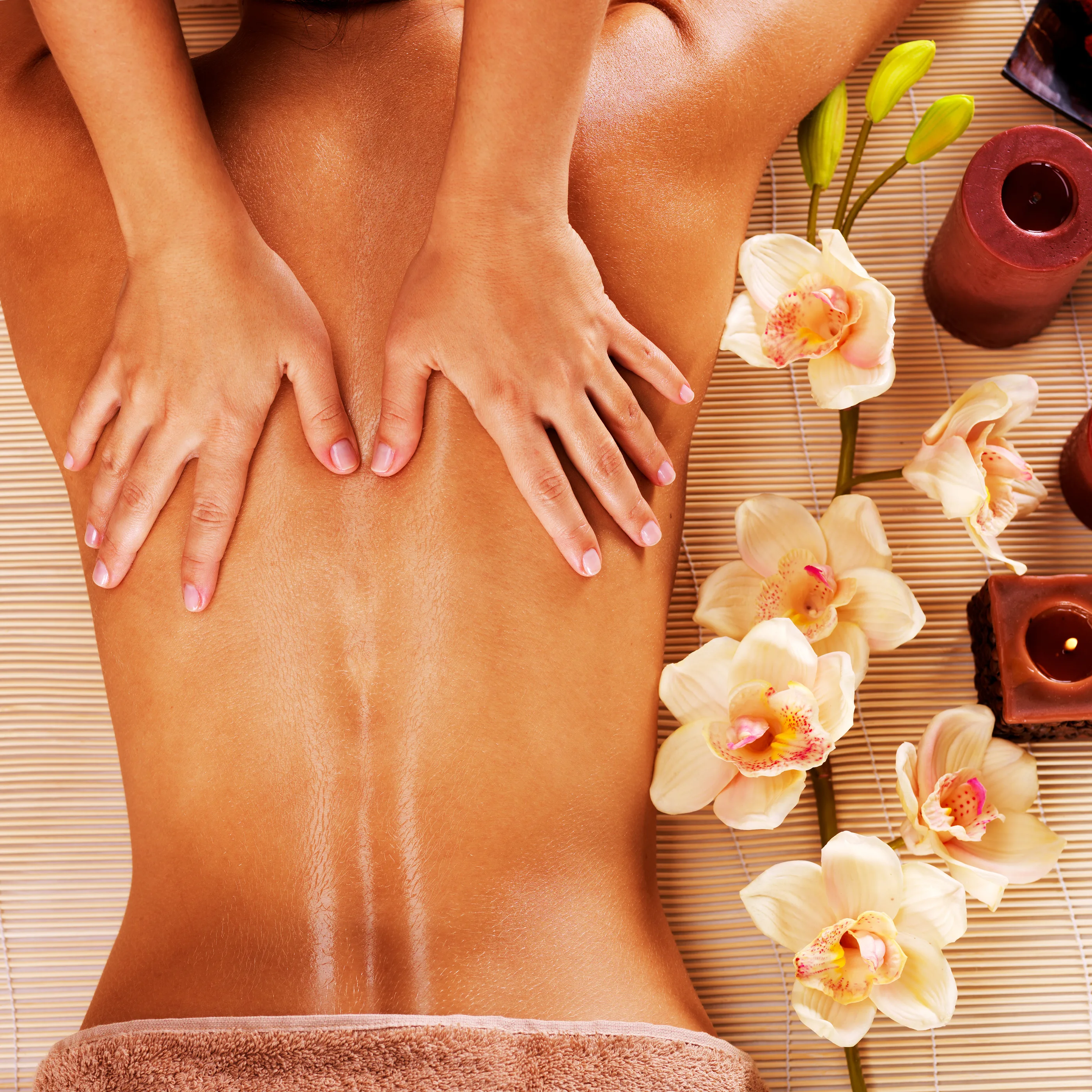 Overhead view of a back massage — therapist's hands on an oiled back, surrounded by cream orchids and lit candles